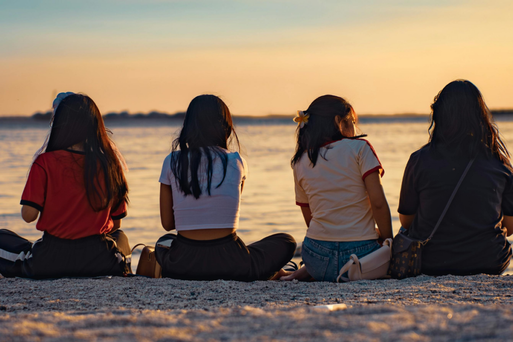 Four friends sit on the beach facing the ocean at sunset