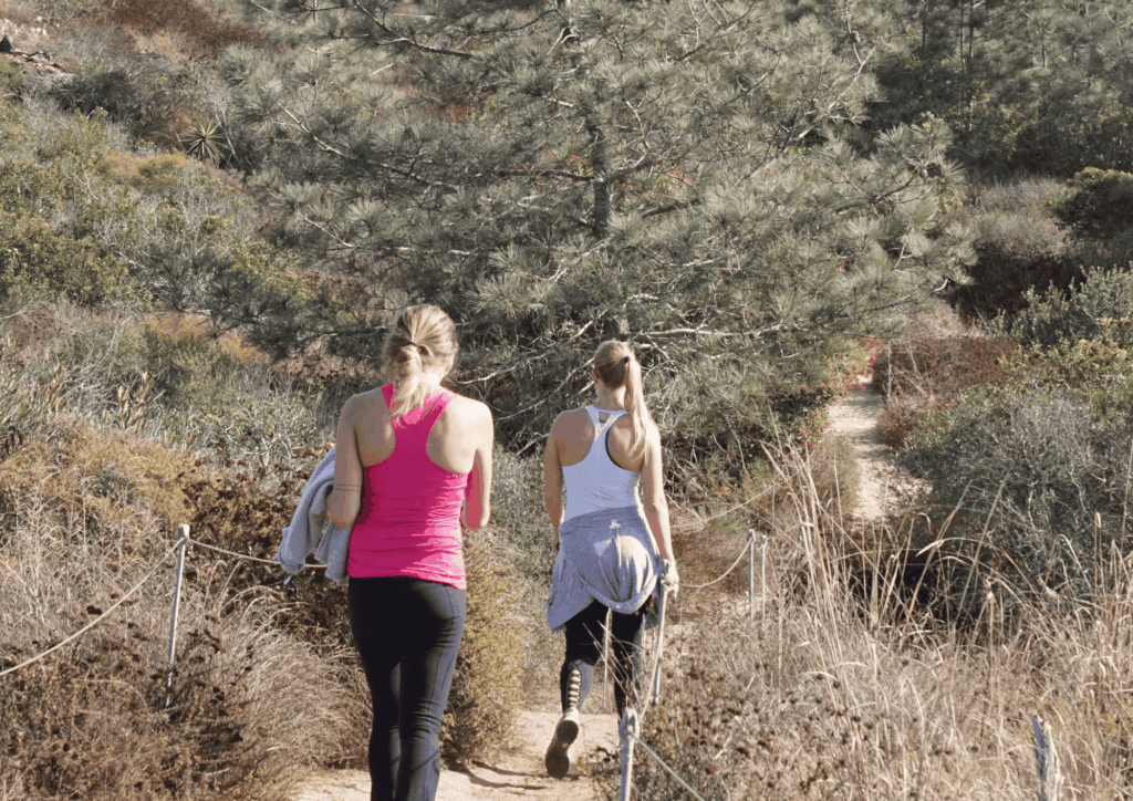Two women walking along a narrow hiking trail through coastal vegetation
