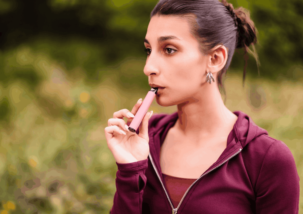 Woman in a burgundy jacket using a pink vape outdoors